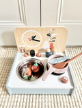 Wooden toy kitchen set with pots, pans, food, and utensils, laid out on a textured rug for realistic play.