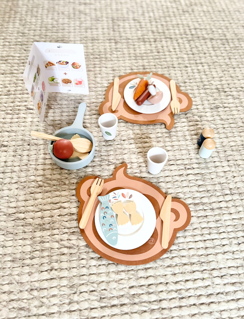 A wooden play dining set arranged on a rug, featuring plates, cutlery, food, and cups, with an instruction booklet.