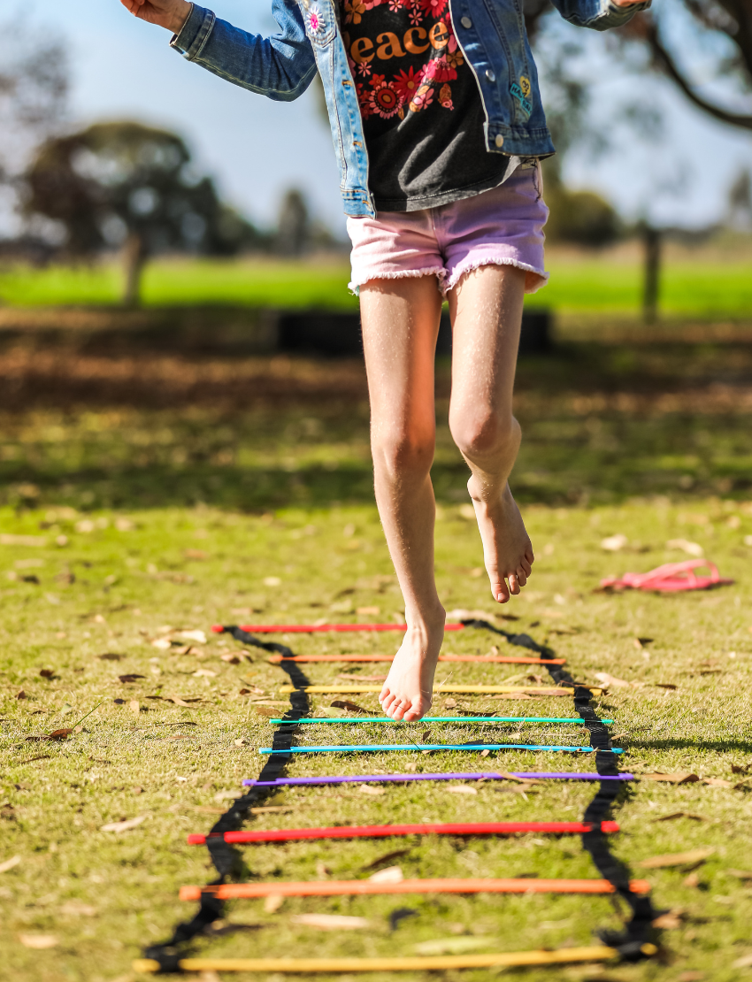 Child's feet jumping through a colorful agility ladder on grass in a park.