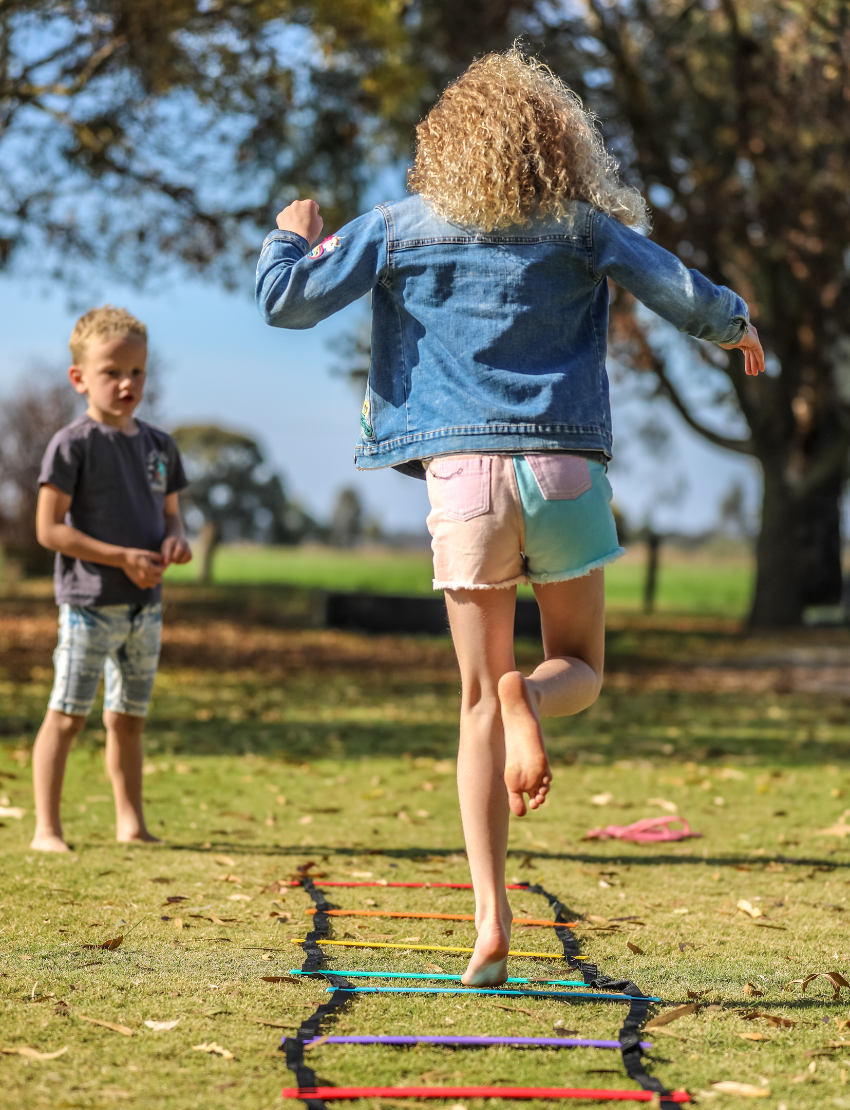 Girl with curly blonde hair, viewed from behind, jumps through an agility ladder in a park. Boy watches.