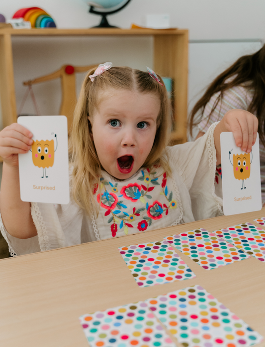 A young child with pigtails, looking surprised, holds a flashcard from The Play Way's "Emotions Part 1: Naming our Feelings Program," featuring a surprised face. She sits at a table with colorful polka-dot cards, and a background shelf displays toys and a globe.