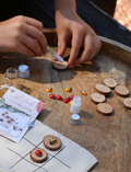 Hands assembling DIY Tic-Tac-Toe game pieces: painting wooden discs and placing ladybug/bee charms.