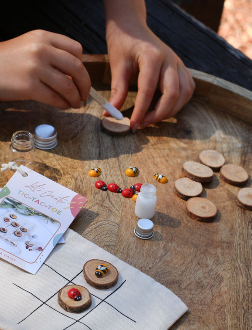 Hands assembling DIY Tic-Tac-Toe game pieces: painting wooden discs and placing ladybug/bee charms.
