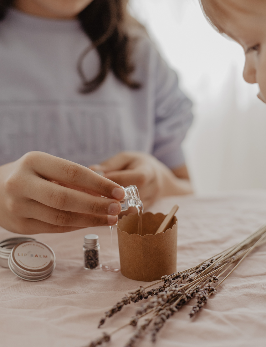 Children making lavender lip balm, pouring oil into a craft cup with dried lavender flowers and ingredients nearby.