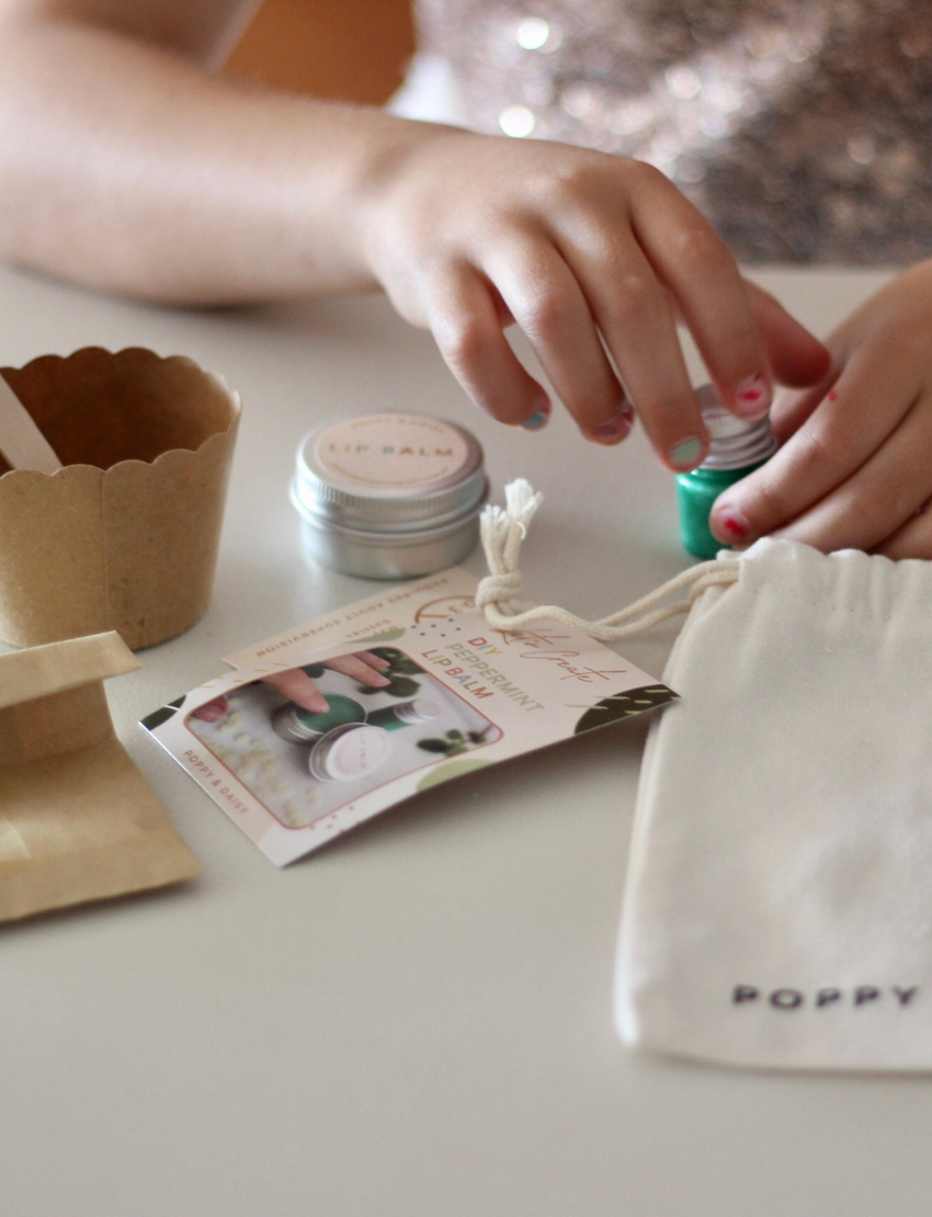 Children's hands working with a DIY Peppermint Lip Balm kit, using lip balm tins and green liquid.