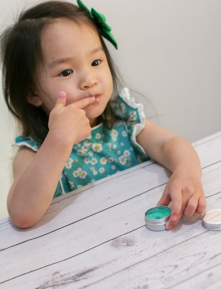 Little girl with painted nails tasting DIY peppermint lip balm, with a tin and green liquid nearby.