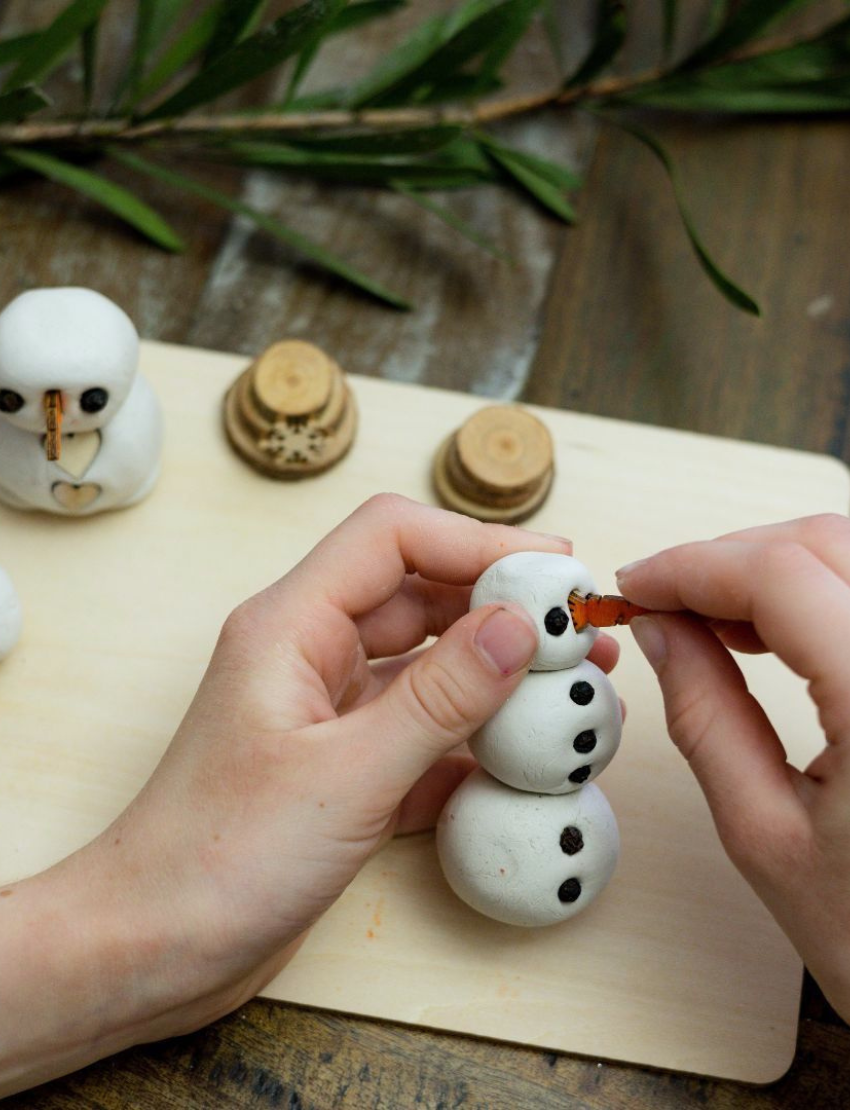 Hands adding an orange carrot nose to a small DIY clay snowman with black bead eyes.