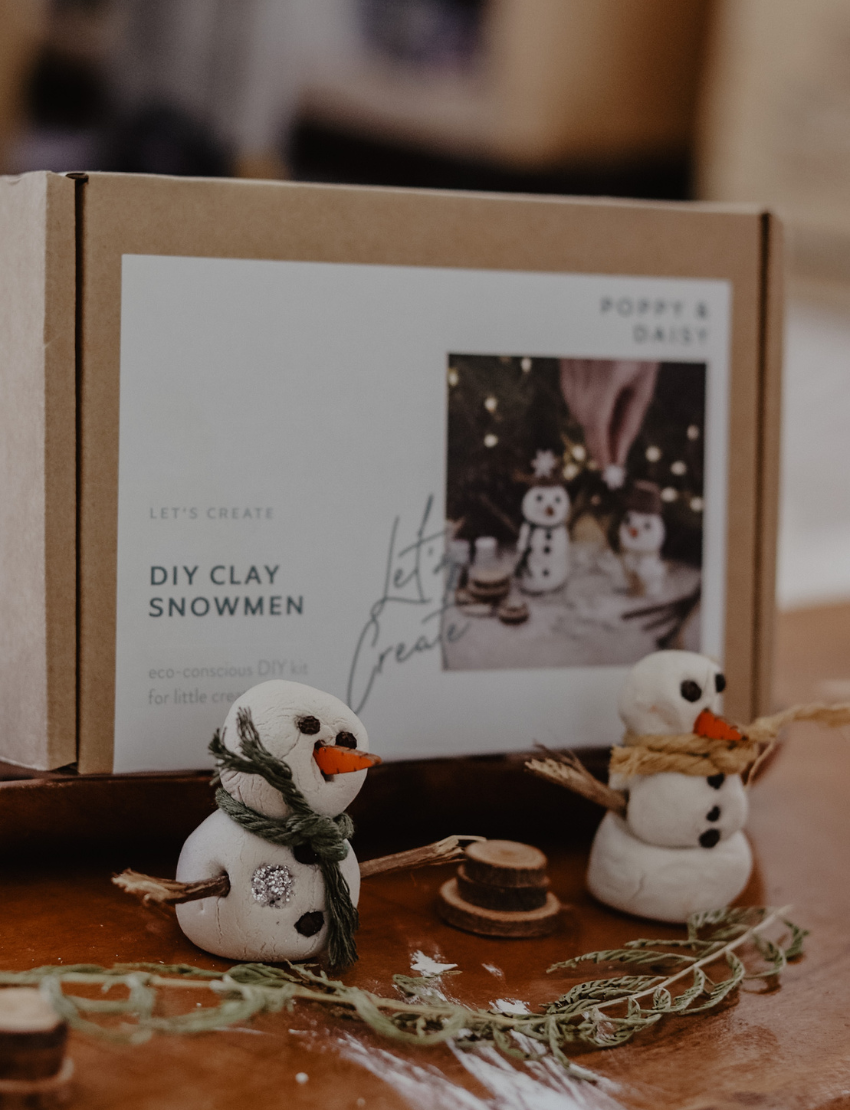 DIY Clay Snowmen kit contents with finished snowmen, box, and crafting supplies on a wooden surface.