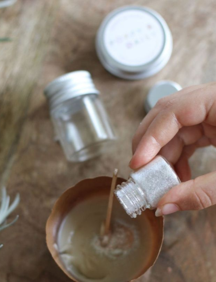 Hand pouring silver glitter from a small bottle into a paper cup with a wooden stick.