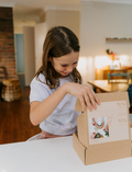 Happy girl opening a Poppy & Daisy Macrame Rainbow DIY kit box, showing the contents inside.
