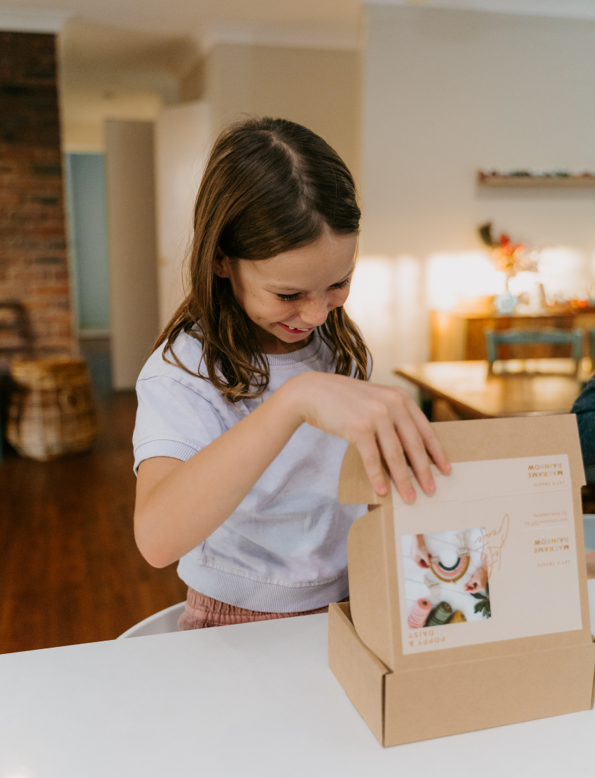 Happy girl opening a Poppy & Daisy Macrame Rainbow DIY kit box, showing the contents inside.
