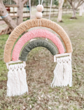 A colorful macrame rainbow decoration with tassels, made from yellow, pink, and green yarn, hanging outdoors.
