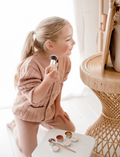 Young girl with blonde ponytail, wearing pink cardigan, applying blush with a brush. Mineral makeup kit nearby.