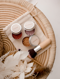 Poppy & Daisy mineral makeup kit: rose, shimmer, bronze powders, brush, and linen pouch on a rattan table.