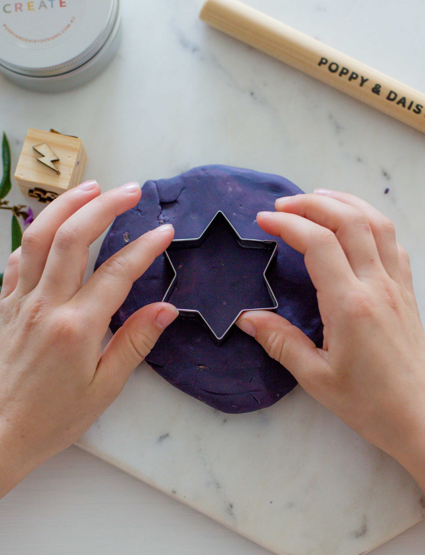 Hands using a star cookie cutter on purple playdough, with wooden stamps and Poppy & Daisy tools.