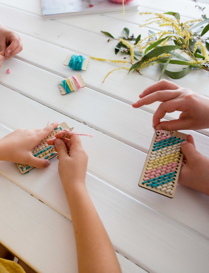 Kids' hands working on DIY embroidered bookmarks, adding colorful yarn to wooden boards for crafting.