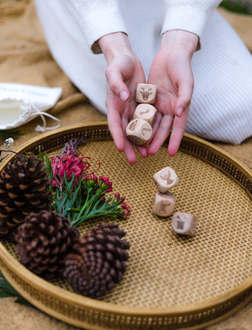 Hands holding wooden story cubes with Christmas icons, surrounded by pinecones and festive foliage.