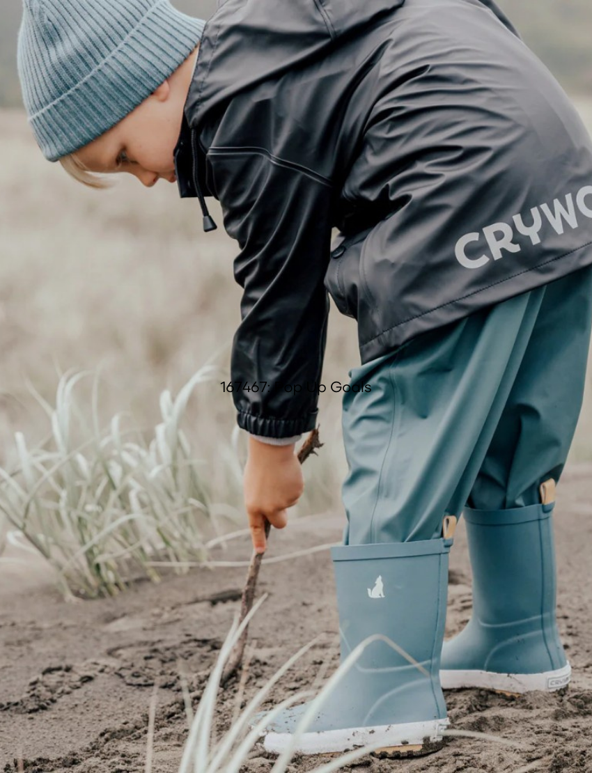 A boy in a black rain jacket and blue rain boots examines a stick on a sandy beach, showing the jacket's 'CRYWOLF' logo.