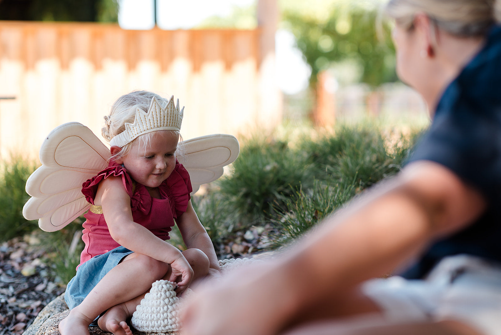 A little girl dressed as a fairy with wings and a crown sits on a rock, interacting with someone out of frame.