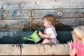 Two children play with watering cans near an outdoor water trough and wooden taps.