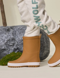 Child's legs wearing tan rain boots and gray sweatpants with 'CRYWOLF' logo, walking near rocks and moss.