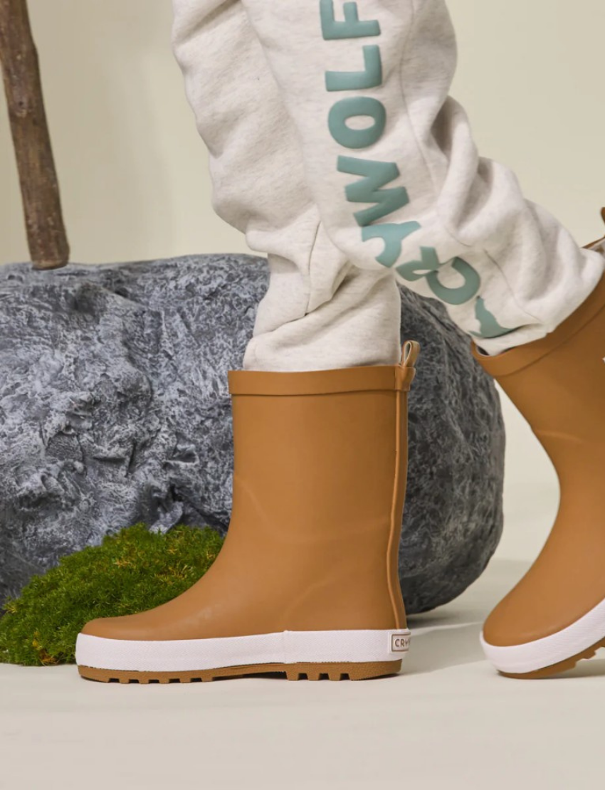 Child's legs wearing tan rain boots and gray sweatpants with 'CRYWOLF' logo, walking near rocks and moss.