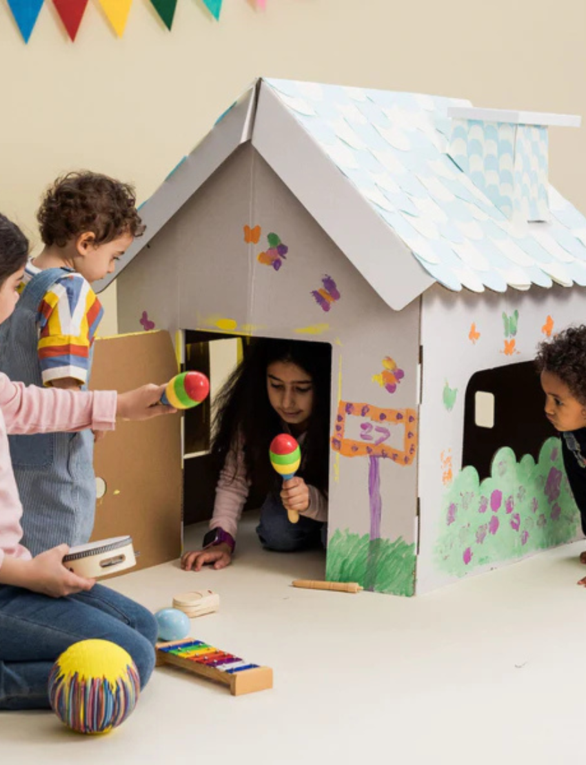 Happy kids playing around a decorated cardboard playhouse. One child peeks inside with maracas, others engage in creative play.