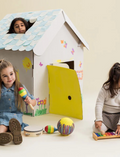 Three happy kids playing with a decorated cardboard playhouse. One peers from the roof, another plays maracas, third plays xylophone.