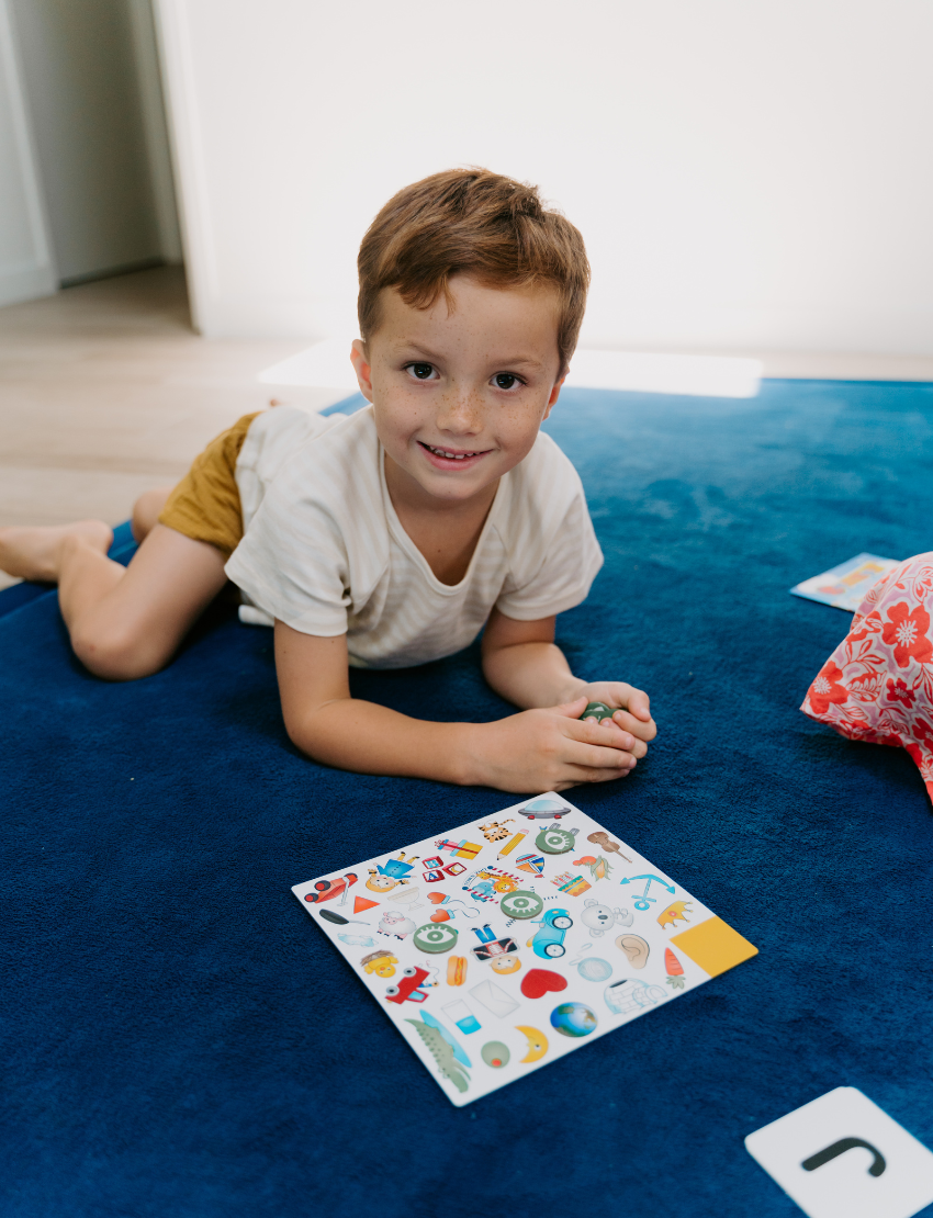 A teal box labeled "Sound Awareness Program" by Therapy At Home holds games, books, spot markers, and activity cards for pre-literacy skills, with some items inside the box and others floating above colorful shapes.