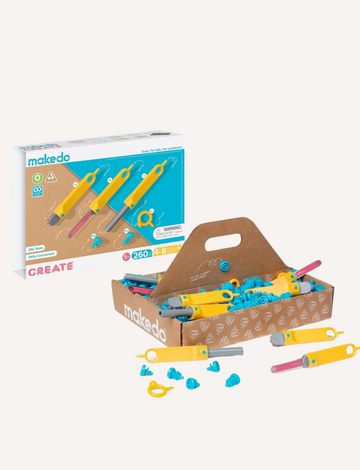 A hand holding the Create Kit's yellow and blue rotary cutter by makedo is shown cutting into brown cardboard against a plain light blue background.