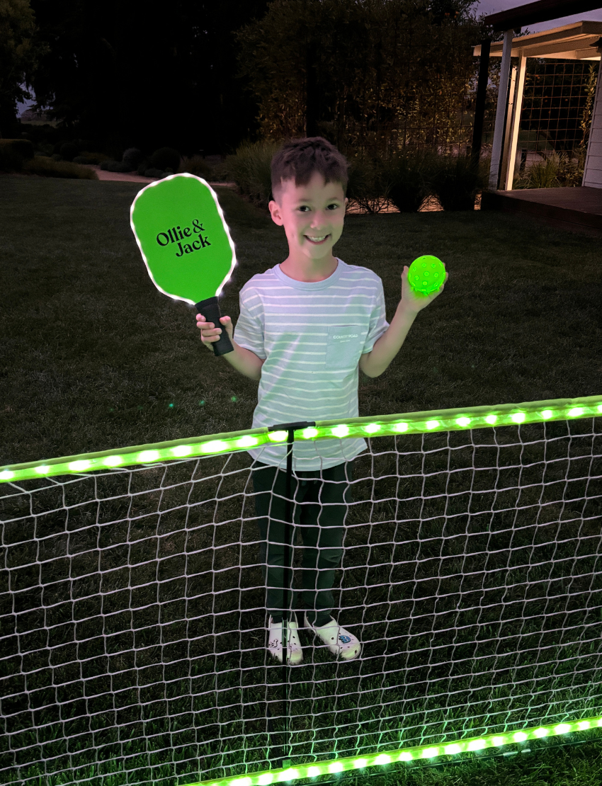 Two kids are smiling and posing outdoors with their arms around each other while holding paddles and balls from the Ollie & Jack LED Pickleball Net Set. They are dressed in casual summer clothing and caps, with greenery in the background beneath a sunny sky.