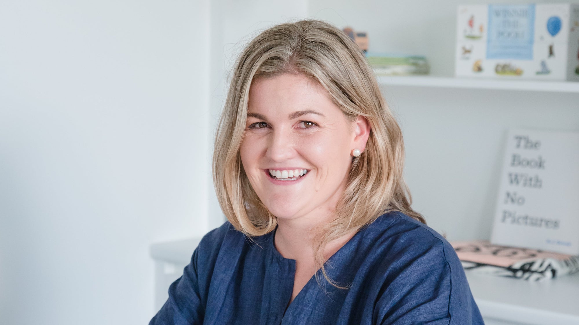 A Kait smiling wearing a navy blue button-up shirt sits at a light wood desk using a laptop in a bright, modern room with white shelves in the background.