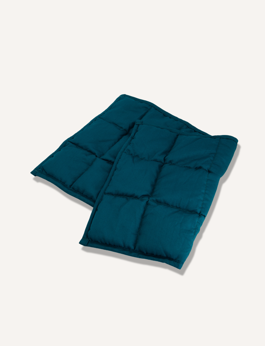 A folded, teal weighted lap pad by The Play Way is displayed on a plain white background, featuring square stitching patterns that create a grid-like appearance.