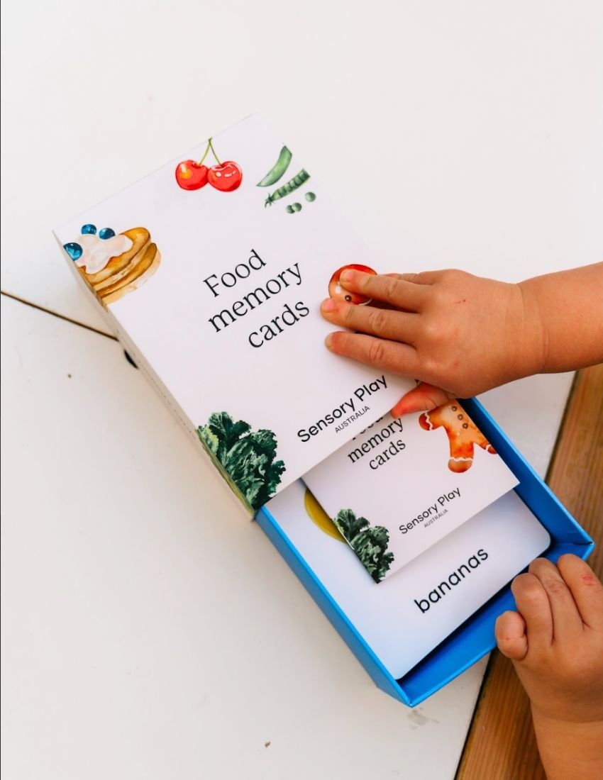 Two Sensory Play Australia "Food Memory Cards Set in Box" packages are shown. The front box displays vibrant illustrations of food items such as broccoli, a gingerbread cookie, and a pie. The back box includes instructions and game information in text and graphics, enhancing the mealtime experience through playful exploration of food.