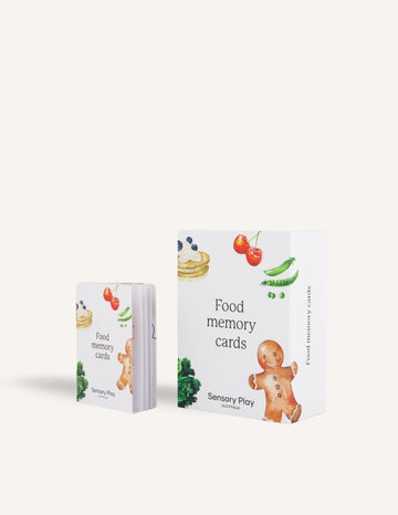 Displayed are two boxes of the "Food Memory Card Set." The larger box on the right features images of a gingerbread person, a cherry, peas, and a dessert. The smaller box on the left displays similar food graphics. Perfect for food exploration, this set is by "Sensory Play Australia.