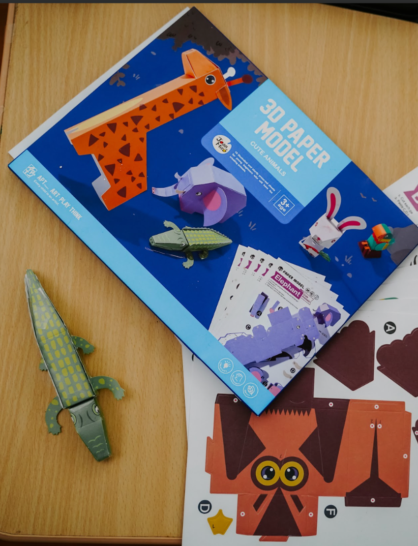 A teal box is open with four colorful "Fine Motor Practice Craft Kits" by The Play Way protruding from it. These kits, ideal for creative play and developing fine motor skills, are labeled "Amazing Origami," "3D Paper Model," "Dream Sand Art," and "Liquid Chalks." Each kit features playful illustrations and vibrant colors.