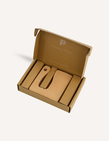 An open brown cardboard box with minimalistic packaging contains the Chop It Training Set from Preppit, featuring a set of wooden items. Inside, you'll find a flat rectangular wooden piece and two other uniquely shaped wooden pieces, all neatly arranged alongside a wavy veggie cutter. The inside of the lid includes the text "Welcome to Preppi.