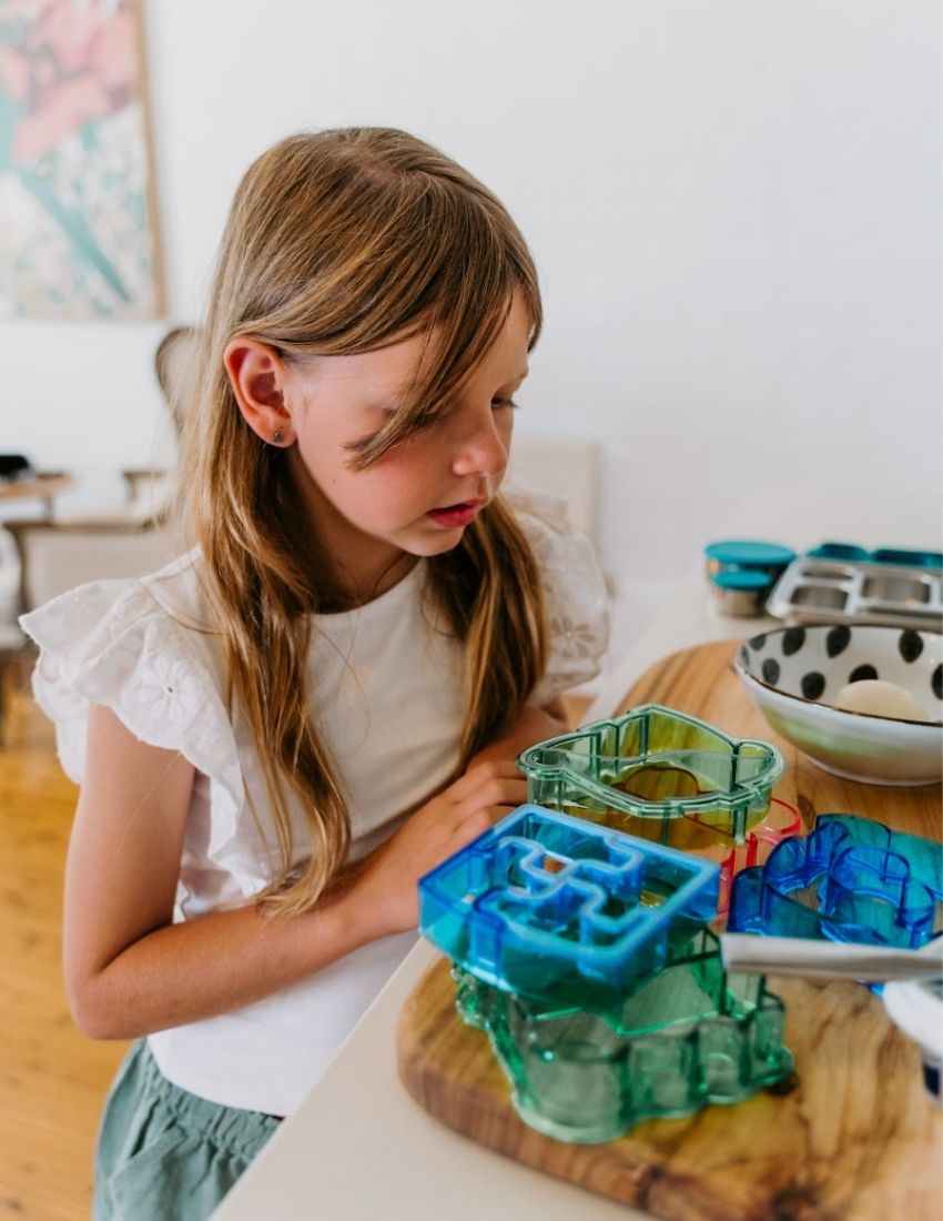 A blue dolphin-shaped sandwich cutter sits in front of a small brown and white box labeled "Lunch Buddy Sandwich Cutters Set" by The Play Way. The box, adorned with wave patterns on the sides, promises fun sandwich shapes designed to delight even the pickiest eaters.