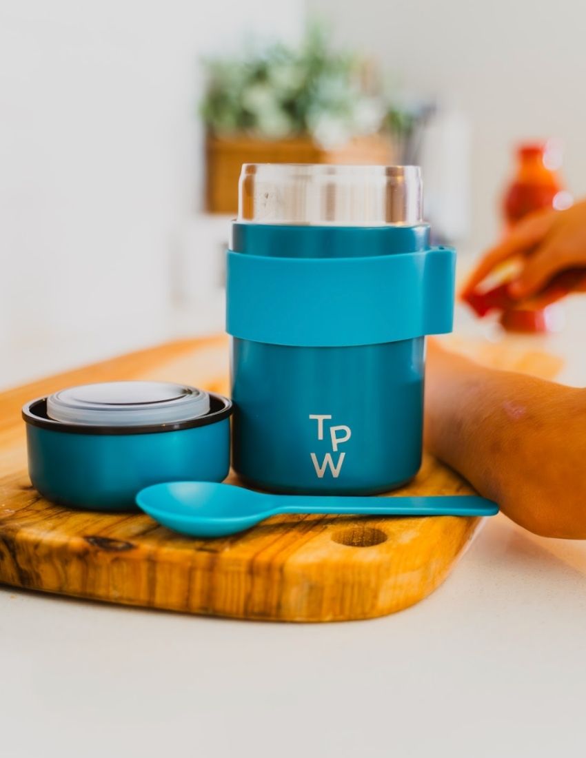 A blue insulated food jar from The Play Way with the initials "T P W" printed on the front. The Lunch Buddy Insulated Container Set features a matching blue silicone band around its midsection that holds a blue spoon, along with a leak-proof lid. The background is plain white.