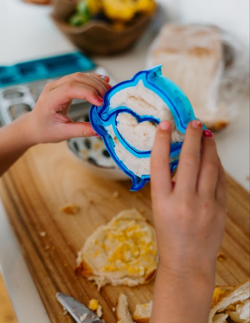 A blue dolphin-shaped sandwich cutter sits in front of a small brown and white box labeled "Lunch Buddy Sandwich Cutters Set" by The Play Way. The box, adorned with wave patterns on the sides, promises fun sandwich shapes designed to delight even the pickiest eaters.