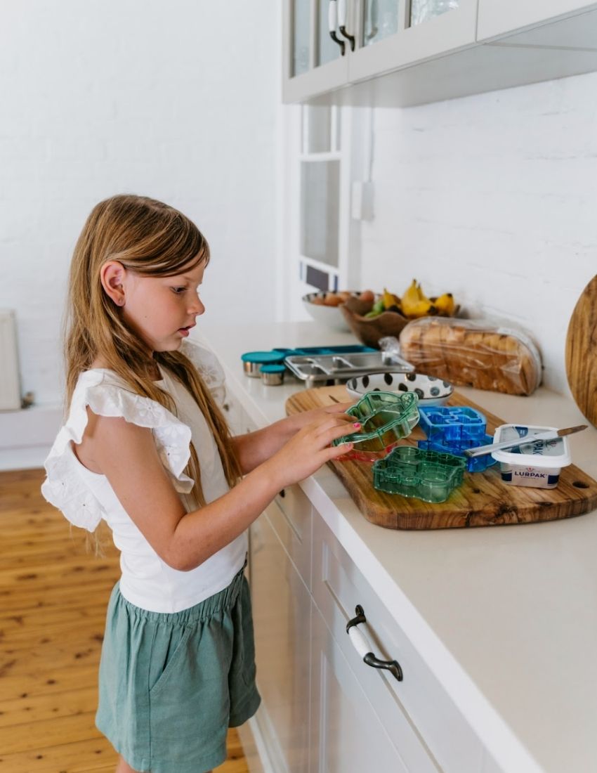 A blue dolphin-shaped sandwich cutter sits in front of a small brown and white box labeled "Lunch Buddy Sandwich Cutters Set" by The Play Way. The box, adorned with wave patterns on the sides, promises fun sandwich shapes designed to delight even the pickiest eaters.