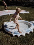 A cardboard box with an open lid contains a Sprinkler Mat package from The Play Way's Sensory Regulation Kit. The package displays a round mat with colorful sections. Two cards are partially placed inside the box, showing a sensory play card and a colored rainbow icon to support emotional regulation activities.