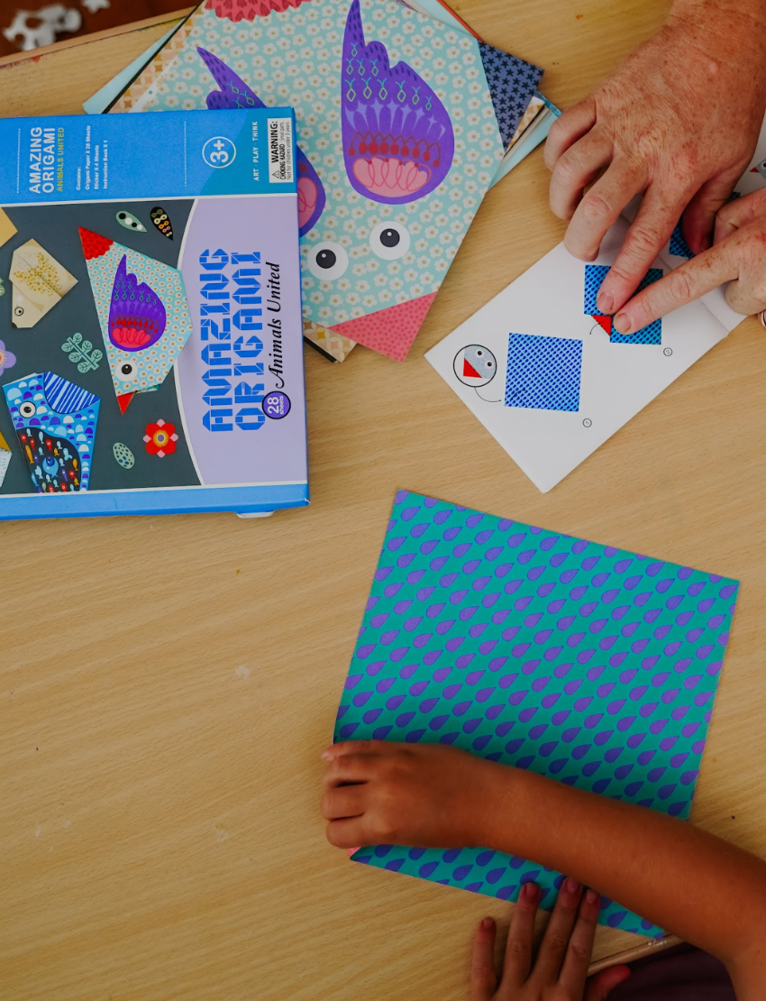 A teal box is open with four colorful "Fine Motor Practice Craft Kits" by The Play Way protruding from it. These kits, ideal for creative play and developing fine motor skills, are labeled "Amazing Origami," "3D Paper Model," "Dream Sand Art," and "Liquid Chalks." Each kit features playful illustrations and vibrant colors.