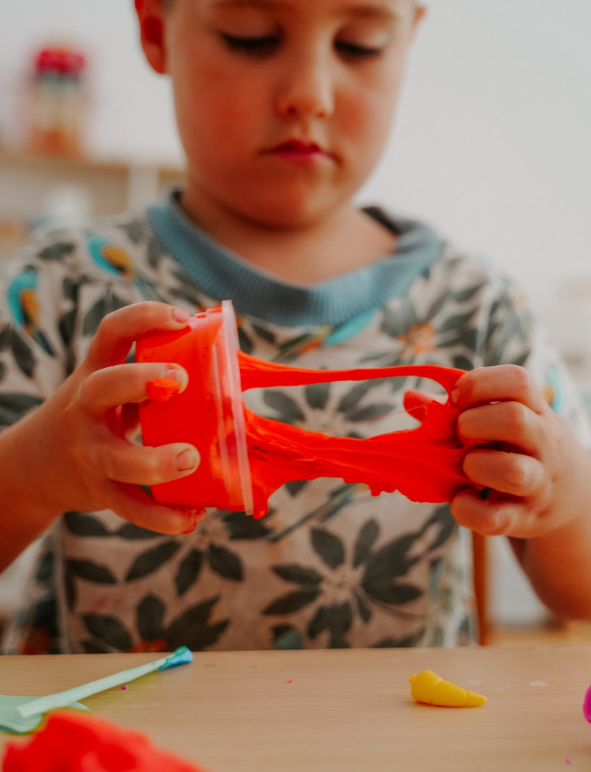 A box of the Fine Motor Practice: Playdough Kit by The Play Way, containing colorful playdough tubes and wooden stamps with various designs for creative expression, along with a booklet titled "Playdough Stamps." The box is adorned with abstract shapes and partially opened to reveal these sensory engagement tools, which are designed to enhance fine motor skills.