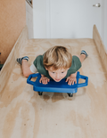 A child with blond hair is on The Play Way's Scooter Board, sliding down a wooden ramp. Wearing a green shirt and striped socks, the child grips the board handles excitedly, enhancing motor skills in this playful activity.