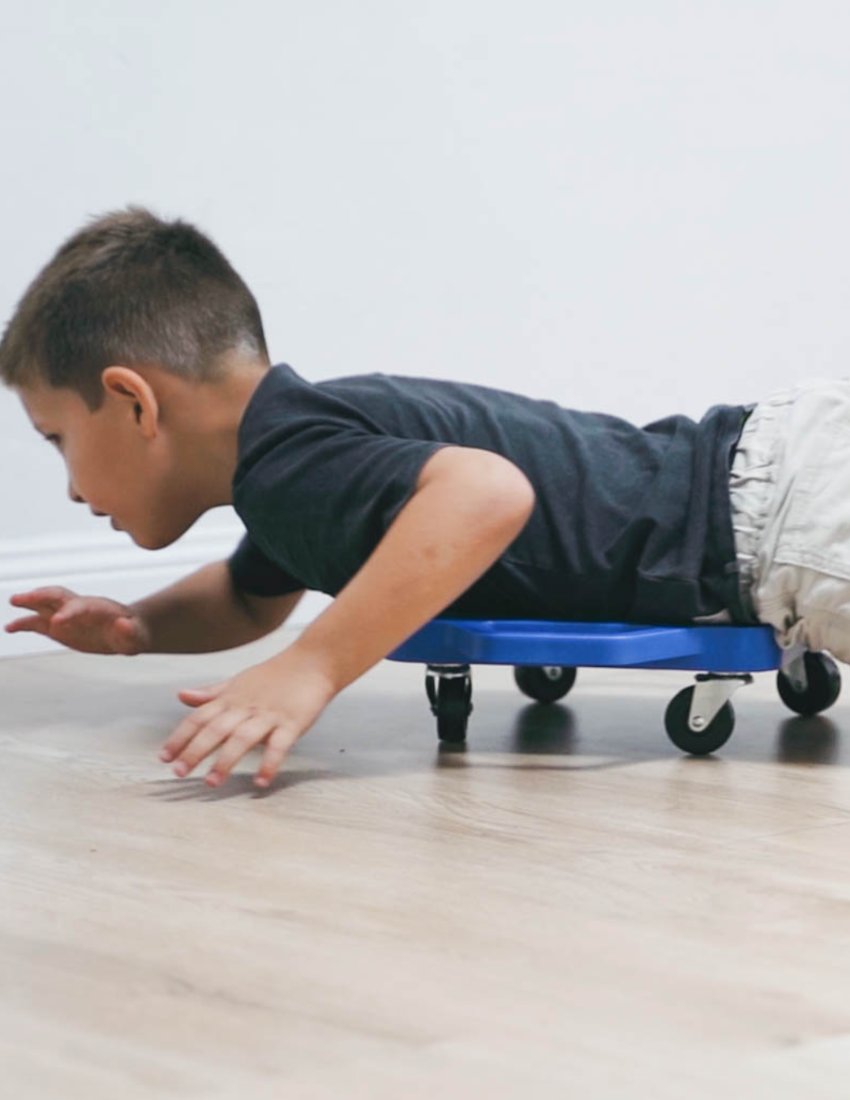 A child with blond hair is on The Play Way's Scooter Board, sliding down a wooden ramp. Wearing a green shirt and striped socks, the child grips the board handles excitedly, enhancing motor skills in this playful activity.