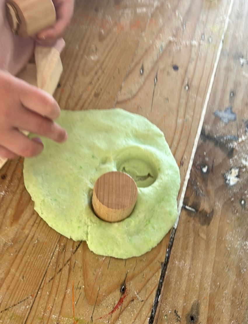 A box of the Fine Motor Practice: Playdough Kit by The Play Way, containing colorful playdough tubes and wooden stamps with various designs for creative expression, along with a booklet titled "Playdough Stamps." The box is adorned with abstract shapes and partially opened to reveal these sensory engagement tools, which are designed to enhance fine motor skills.