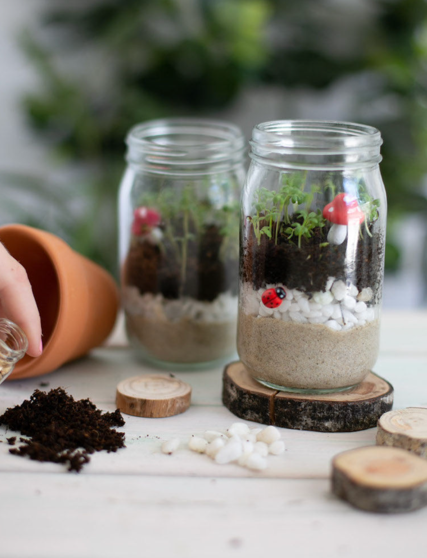 A photo of an open box displaying a variety of items from The Play Way's Gardening and Handwriting Kit, including a book titled "Grow," a drawstring bag, a pack of birdseed, a Melitta coffee pack, and crafting materials like a mushroom-shaped item and wooden spoon for fine motor development.