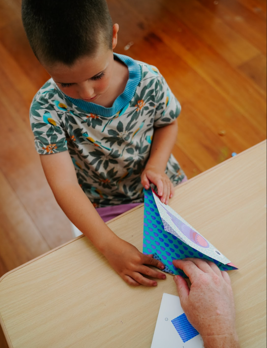 A teal box is open with four colorful "Fine Motor Practice Craft Kits" by The Play Way protruding from it. These kits, ideal for creative play and developing fine motor skills, are labeled "Amazing Origami," "3D Paper Model," "Dream Sand Art," and "Liquid Chalks." Each kit features playful illustrations and vibrant colors.