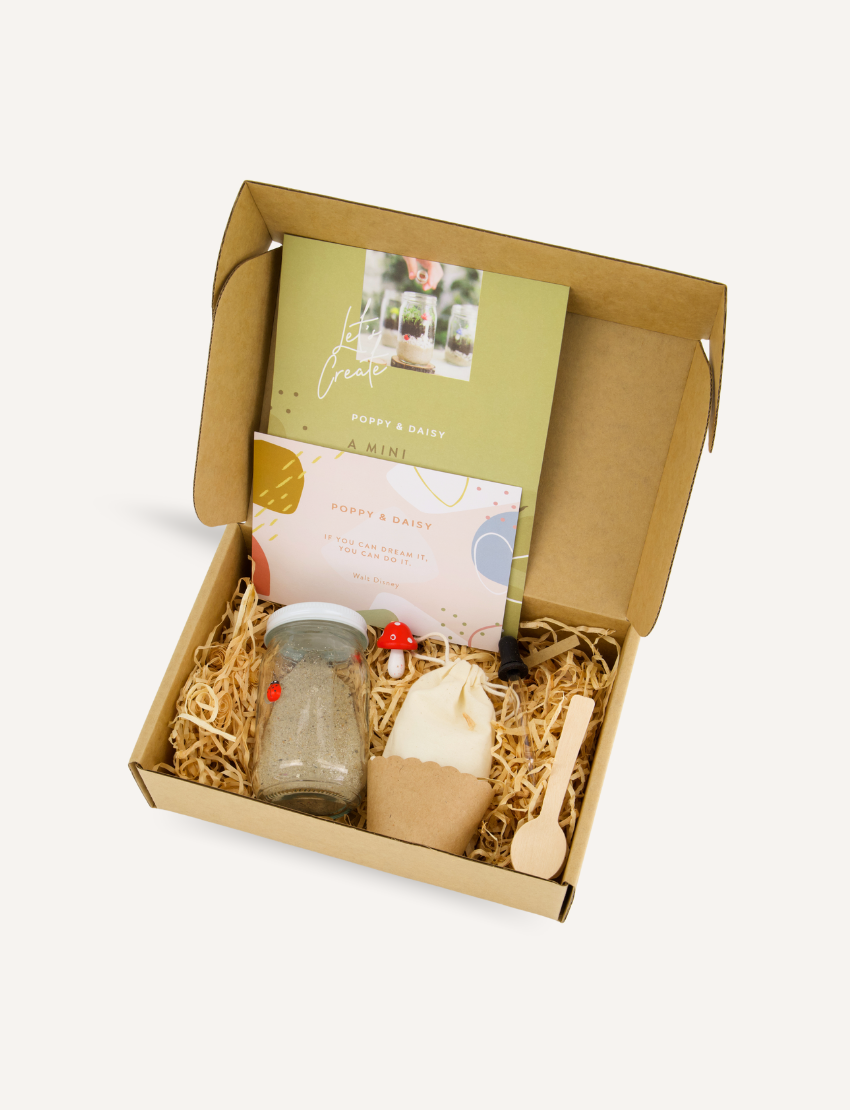 A photo of an open box displaying a variety of items from The Play Way's Gardening and Handwriting Kit, including a book titled "Grow," a drawstring bag, a pack of birdseed, a Melitta coffee pack, and crafting materials like a mushroom-shaped item and wooden spoon for fine motor development.